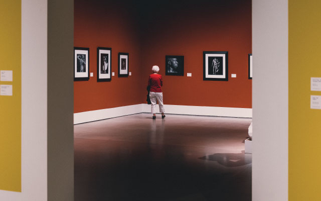 Woman in red sweater observing black and white photographs in an art gallery with a vibrant orange wall, reflecting Bentonville's cultural scene.